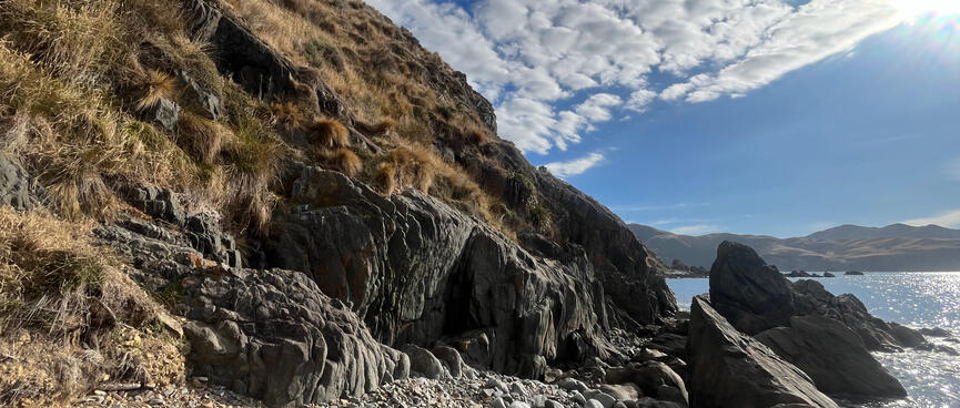 The gap between a steep tussock-clad hill and angled rocks rising from the sea.