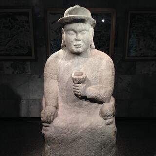 A stone man wears a bowler hat and holds a cup in one hand.