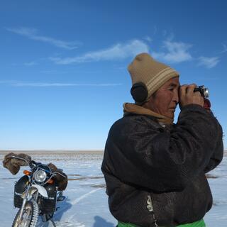 A man with a black jacket, brown hat and ear muffs surveys the landscape with my monocular. His motorcycle is parked behind him.