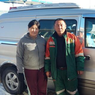 A woman in street clothes and a man in overalls stand in front of their van.