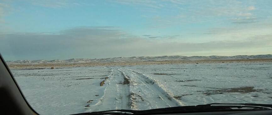 View out the windscreen to tyre tracks through the snow, intermittently revealling patches of brown sand.