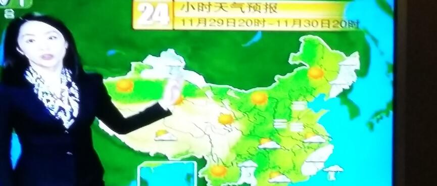 Still frame of a TV weather programme showing a Chinese woman dressed in a suit and floral blouse pointing to sun and cloud icons on a map of China.