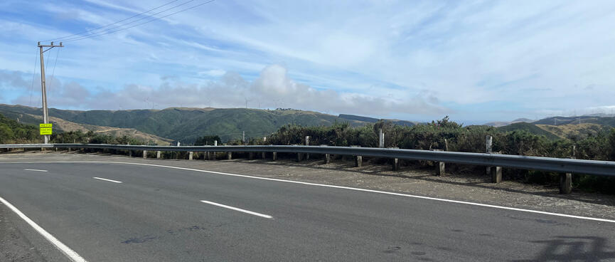 On the side of a two lane road, wind turbines are visible on distant hill tops.