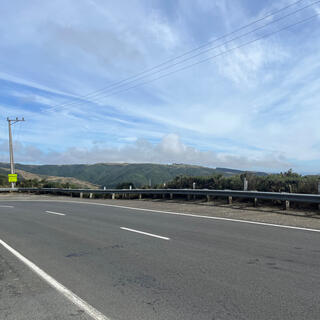 On the side of a two lane road, wind turbines are visible on distant hill tops.