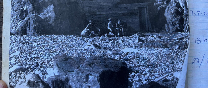 Three men sit outside the cave, John Magurk at left. Someone has written ʼ1960s?ʼ.