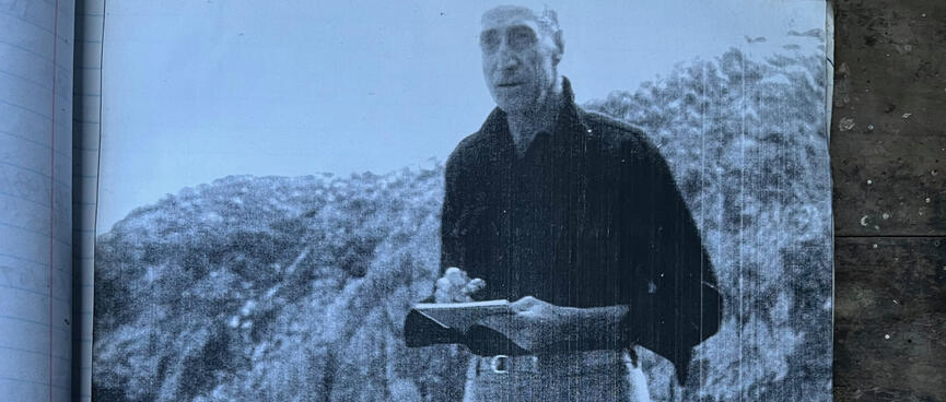A tall man wearing a dark shirt and baggy pants holds a sketch pad.