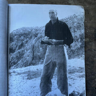 A tall man wearing a dark shirt and baggy pants holds a sketch pad.