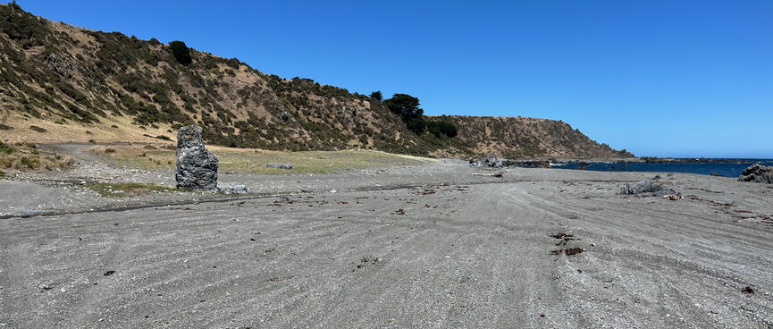A vertical slab of stone on the left of a wide shingle beach.