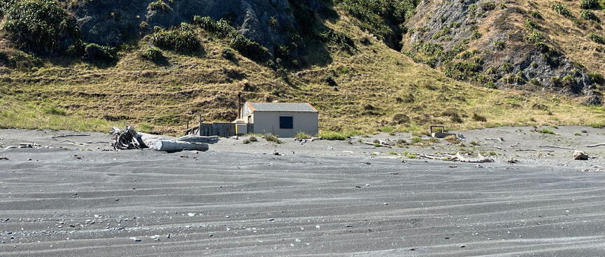 A simple house at the foot of grassed hills on the edge of a shingle beach.