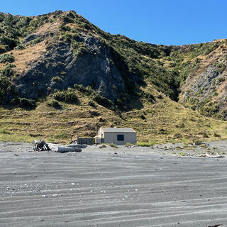A simple house at the foot of grassed hills on the edge of a shingle beach.