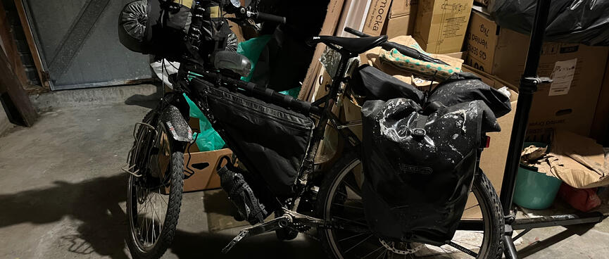My loaded touring bike leans against a pile of boxes in a dark garage.