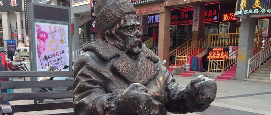 A bronze statue of a seated man wearing a hat, trench coat and boots, and holding his hands up as if to hold reins.