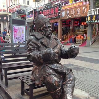 A bronze statue of a seated man wearing a hat, trench coat and boots, and holding his hands up as if to hold reins.