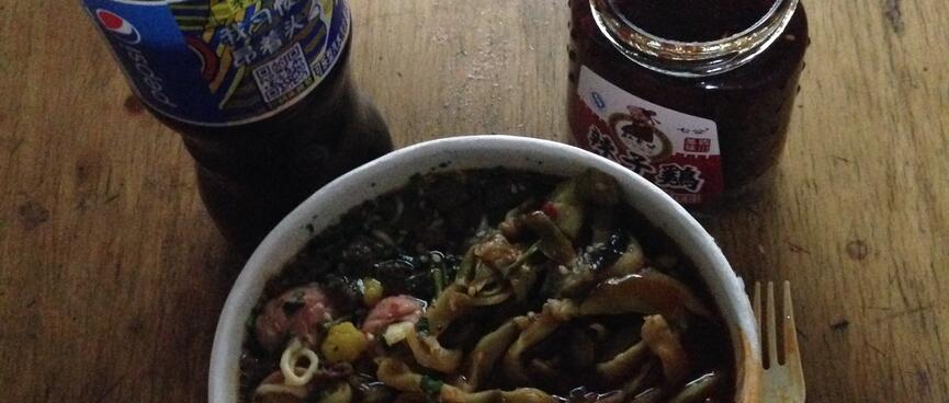 A container of cooked instant noodles, with added sausage and spicy greens, a Pepsi and a jar of chilli.