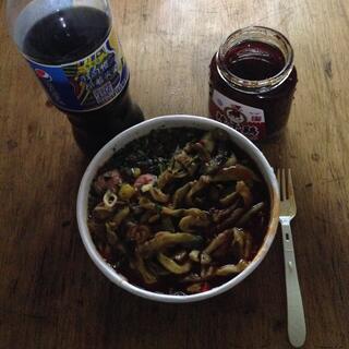 A container of cooked instant noodles, with added sausage and spicy greens, a Pepsi and a jar of chilli.
