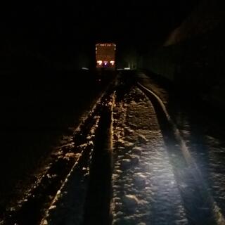 A car's headlights illuminate the snowy road ahead.