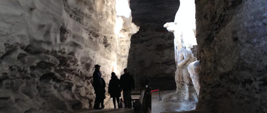 A tall narrow cavern where people view a huge carving bathed in a beam of light.