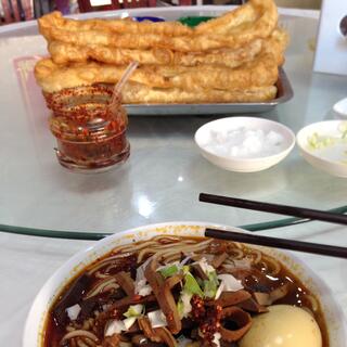A bowl of noodle soup and a metal tray stacked with long golden fritters.