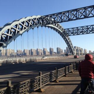 Zhihong rides under curved steel beams at the crest of a bridge, where many high rises are visible in the distance.
