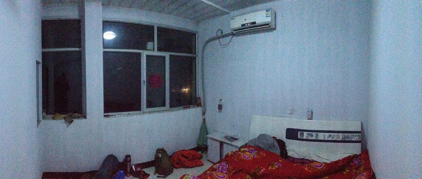 A room with white walls, a heatpump, windows with no curtains and a double bed with a red bedspread.