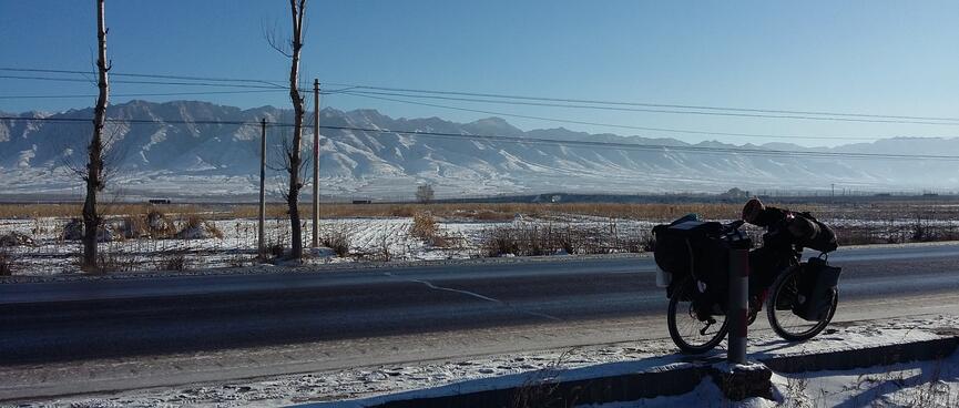 My bicycle leans against a post, snowy fields and a long mountain range appear in the background.