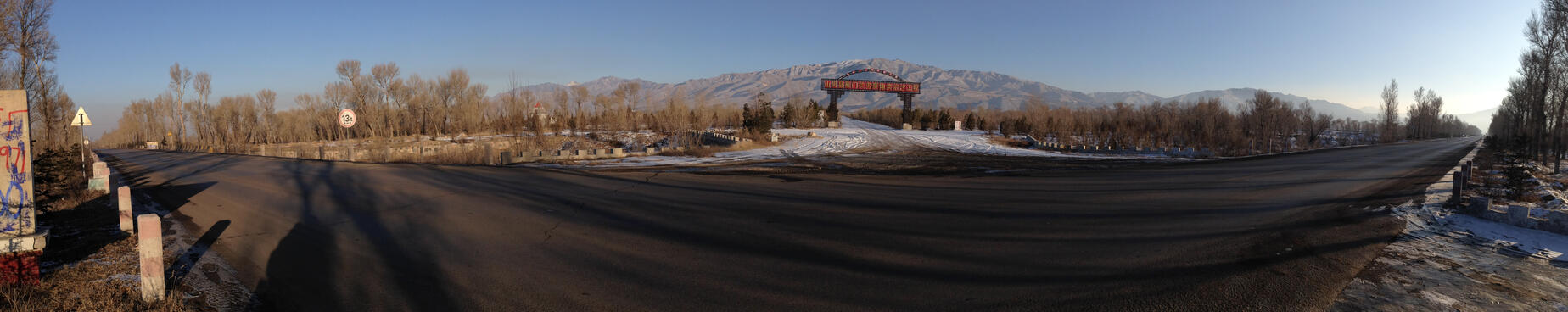 A panoramic shot of a long road lined with trees. A large archway spans a side road heading towards tall mountains.