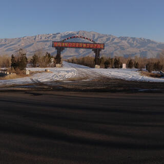 A panoramic shot of a long road lined with trees. A large archway spans a side road heading towards tall mountains.