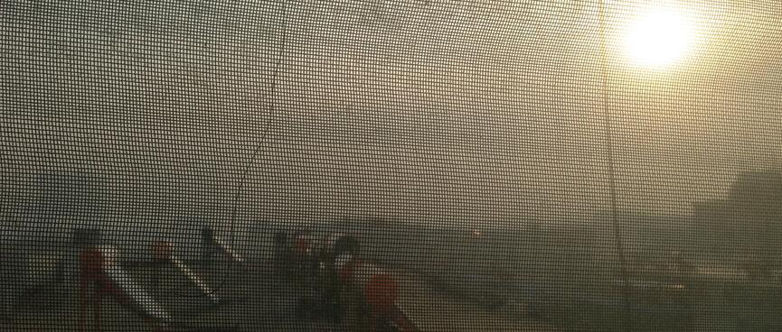 View through sun blinds to a yellow haze surrounding the sun and clouds above solar heating panels on an adjacent rooftop.