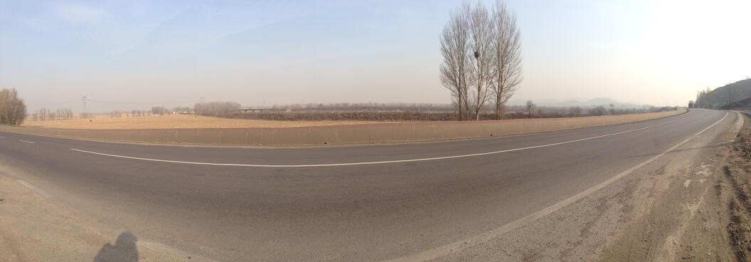 An empty road next to golden fields, with trees and mountains in the distance under a yellowy haze.