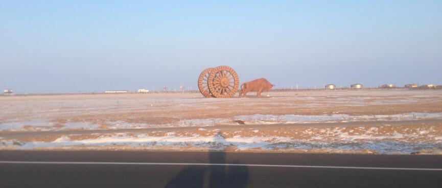 The Troll’s shadow on the road in front of a field containing a metal ox strainingå against two giant wagon wheels.