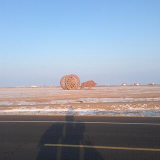 The Troll’s shadow on the road in front of a field containing a metal ox straining against two giant wagon wheels.