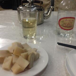 A tea pot and a glass of pale yellow tea, a liquor bottle and a glass of clear liquid, and a white plate containing rectangles of potato.