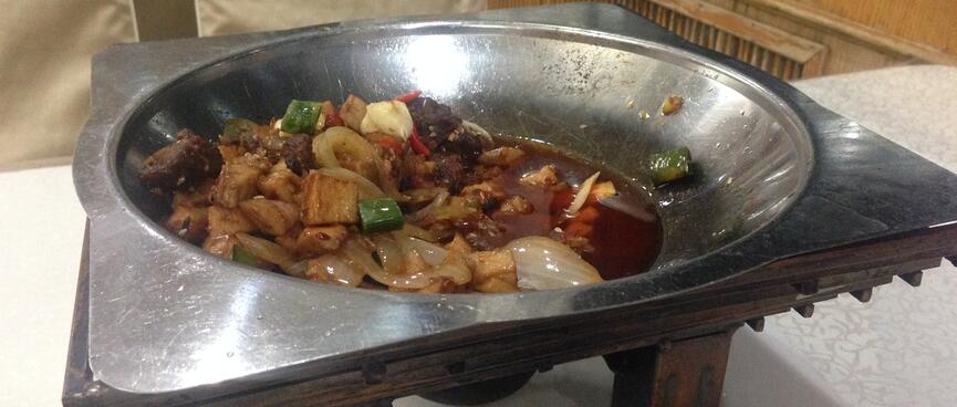 A wide metal heating bowl containing meat and vegetables in an oily red sauce.