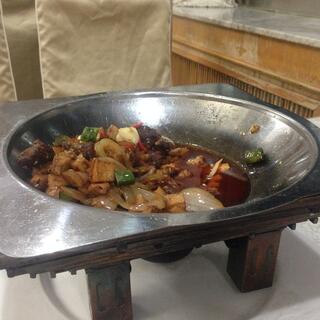A wide metal heating bowl containing meat and vegetables in an oily red sauce.