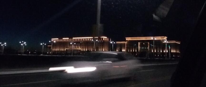 Car headlights streak past a sprawling building illuminated by uplights.
