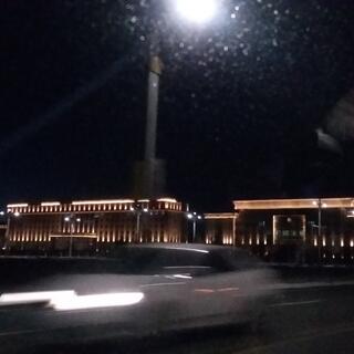 Car headlights streak past a sprawling building illuminated by uplights.