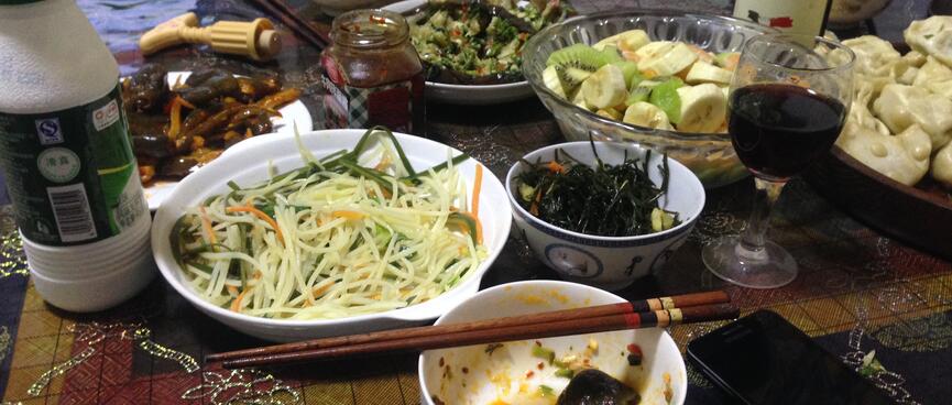 A bottle of wine and serving bowls containing noodles, stir fries, dumplings and fermented vegetables.