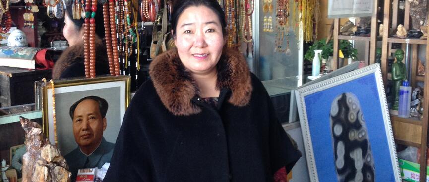 A shopkeeper stands in front of a rack of beaded necklaces and a photo of Chairman Mao.