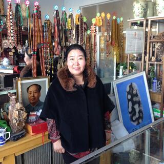A shopkeeper stands in front of a rack of beaded necklaces and a photo of Chairman Mao.