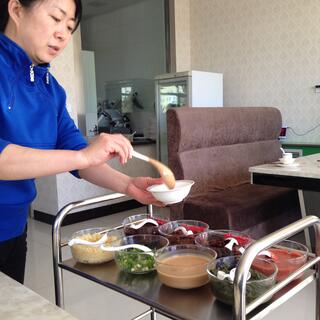A woman in a blue top serves food from a tea trolley filled with bowls of food.