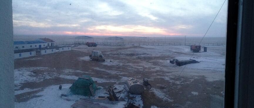 A large fenced dirt yard containing a few scattered vehicles and piles of building materials amongst patches of snow.