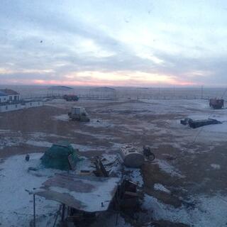 A large fenced dirt yard containing a few scattered vehicles and piles of building materials amongst patches of snow.