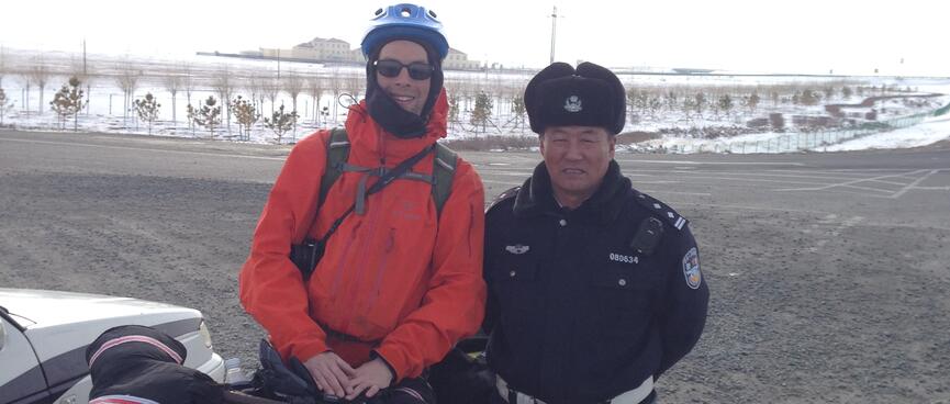 I pose with a policeman who is dressed in black clothes, a black fur hat and a white belt.