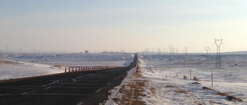 Power pylons on a bank along the right hand side of a road lined with snow and patches of brown dirt.