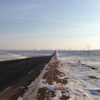 Power pylons on a bank along the right hand side of a road lined with snow and patches of brown dirt.