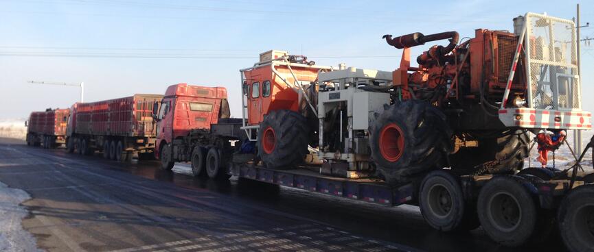 Three trucks parked on the side of the road, the one at the rear is hooked up to a long trailer containing unidentified machinery.