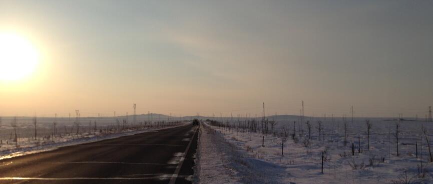 A golden ball hangs low in the sky above a dusky landscape of empty highway and snow covered fields.