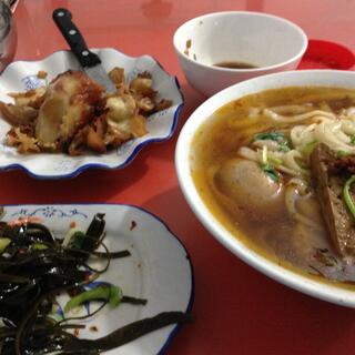 A wide bowl of soup containing thin noodles, meat, egg and thin green vegetables.