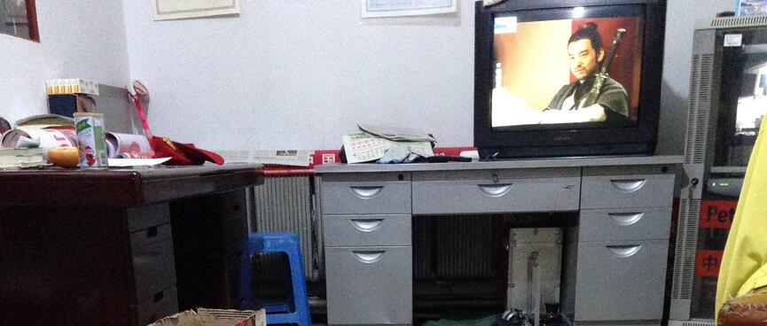 A small white dog sits at my boot clad feet and faces a TV which shows a still frame of an Asian warrior with a sword.