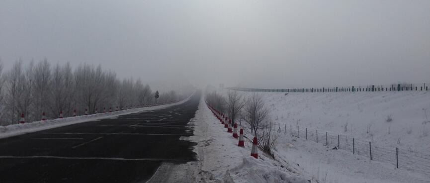 A long highway bordered by trees and snow. The horizon is obscured with mist.